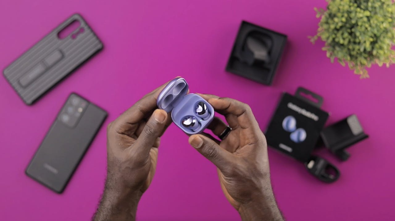 A detailed close-up of hands holding wireless earbuds, surrounded by tech accessories on a vibrant pink background.